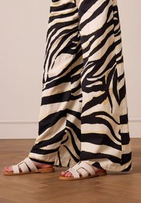 White leather slide sandals with a flat sole featuring crisscross straps. Paired with black and white striped wide-leg pants on a wooden floor.