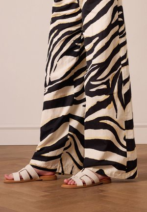Witte leren slip-in sandalen met een platte zool en crisscross banden. Gepaard met zwart-wit gestreepte wijde pantalons op een houten vloer.