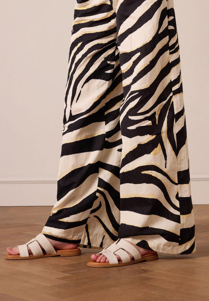 White leather slide sandals with a flat sole featuring crisscross straps. Paired with black and white striped wide-leg pants on a wooden floor.