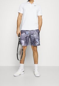 Vit polo skjorta, mönstrade lila shorts med blå detaljer, vita sneakers, håller en tennisracket, mot en neutral bakgrund.