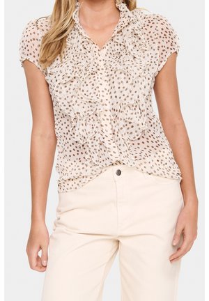 Woman wearing short-sleeve, cream blouse with brown spots and ruffles paired with high-waisted cream pants, standing against white background.