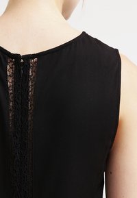 Schwarzes ärmelloses Top mit einer Spitzendetail auf der Rückseite, rundem Ausschnitt und glatter Stoffstruktur. Reißverschlussverschluss im Nacken.
