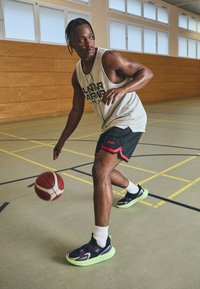 Korvpallur dribblib palli, kandes valget võrksärki, musti lühikesi pükse punaste detailidega ning musti ja rohelisi jooksujalatseid.
