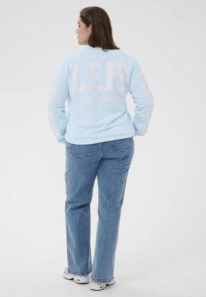 Vrouw die met de rug naar de camera staat, gekleed in een lichtblauwe trui met de tekst "LEIS", blauwe spijkerbroek en witte sneakers, tegen een effen witte achtergrond.