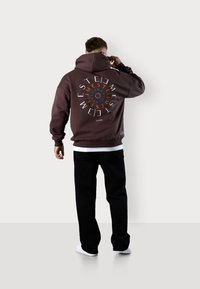 Hoodie marron avec un design graphique au dos, associé à un pantalon noir et des chaussures blanches. Présente une coupe décontractée et des poignets côtelés.