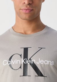 Gaiši pelēks kokvilnas T-krekls ar lielu melnu "K" un baltu "Calvin Klein Jeans" uzrakstu. Klasiska apaļa izgriezuma kakla līnija un mīksta tekstūra.