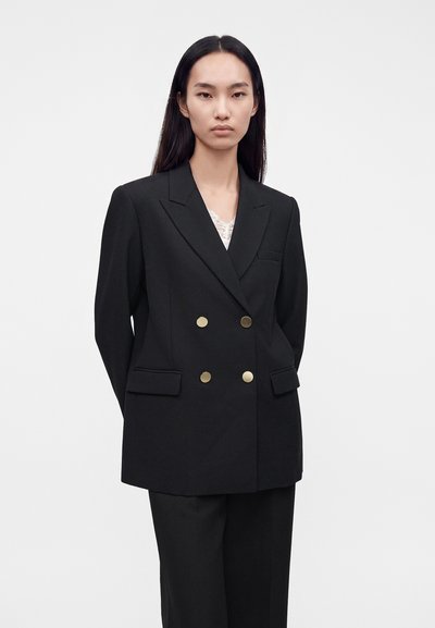 Femme aux longs cheveux foncés portant un blazer noir croisé avec des boutons dorés et un pantalon noir assorti sur un fond clair uni.