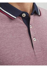 Gros plan sur une personne portant un polo rose texturé avec un col bleu marine comportant des rayures blanches et bordeaux ainsi que des boutons blancs.