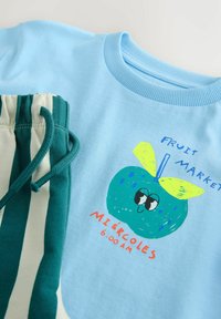 Camisa azul claro con una manzana de dibujos animados en tonos verde azulado con hojas amarillas, texto "Fruit Market" y "Miércoles 6:00 AM", combinada con pantalones a rayas verdes y blancas.