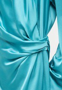 Κοντινό πλάνο λείου, γυαλιστερού τιρκουάζ υφάσματος που είναι μαζεμένο και ντυμένο με μια στριμμένη λεπτομέρεια κόμπου, δημιουργώντας απαλά πτυχώσεις και υφή.