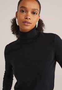 Jeune femme aux cheveux bouclés portant un pull à col roulé noir et de grandes boucles d'oreilles en argent, faisant face à l'avant sur fond neutre.