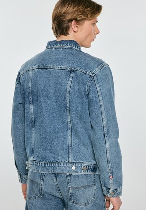 Młody mężczyzna widziany od tyłu, ubrany w jasnoniebieską kurtkę dżinsową i pasujące dżinsy, na neutralnym tle.