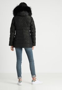 Svart puffjacka med texturerad yta, med en avtagbar svart pälskapuschong, bärs över ljusblå jeans och vita sneakers.