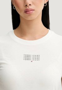 Bílé bavlněné tričko s opakujícím se vzorem textu "TOMMY JEANS". Kulatý výstřih a krátké rukávy, s malým logem v dolní části.