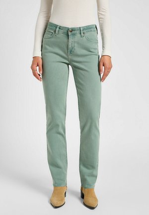 Kobieta ubrana w jasnolistne zielone proste dżinsy, beżowe zamszowe botki do kostki oraz kremowy prążkowany top z długim rękawem, stojąca na jednolitym tle.