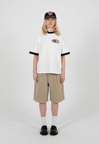 Person iført en hvid oversized T-shirt med et "PMCo"-logo, beige knælange shorts, hvide sokker, sorte sko og en sort kasket med et cirkulært emblem.