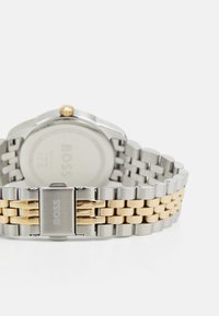 Relógio de pulso em tons de prata e dourado com uma pulseira de elos metálicos, apresentando uma caixa redonda, um botão e o nome da marca gravado no fecho.