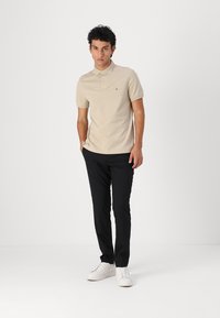 Beige kortærmet polo t-shirt med krave, parret med sorte bukser og hvide sneakers, stående mod en hvid baggrund.