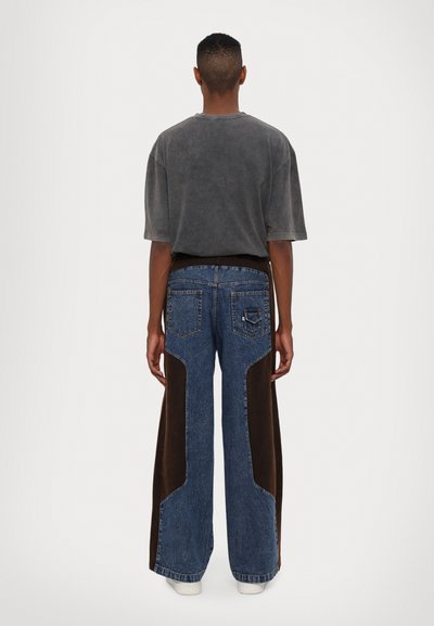 T-shirt gris surdimensionné et jean en denim bleu avec des panneaux latéraux en daim marron, présentant une coupe ample et des poches arrière traditionnelles.