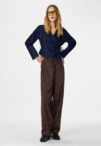 Haut en denim à nouer avec manches longues, associé à un pantalon large à rayures marron. Le mannequin porte des lunettes à verres jaunes et des chaussures noires brillantes.