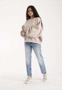 Naine, kellel on pikad lokkis juuksed, kannab üle suuruses beeži värvi pusa, helesiniseid teksaseid ja valgeid tossusid, seisab käed rüpes.