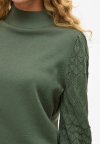 Groene gebreide trui met een hoge kraag en geprofileerde kant mouwen met diamantmotieven. Zacht, lichtgewicht stof met een aansluitend ontwerp.