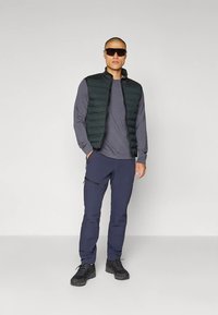 Moški z temnimi sončnimi očali, črno puhasto telovnikom, sivo majico z dolgimi rokavi, modrimi cargo hlačami in črnimi pohodniškimi čevlji stoji ob belem ozadju.