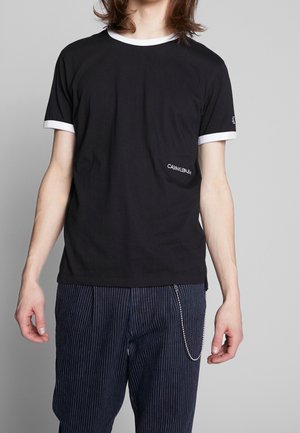 Persoon draagt een zwart Calvin Klein t-shirt met witte rand en marineblauwe gestreepte broek met een zilveren kettingaccessoire.