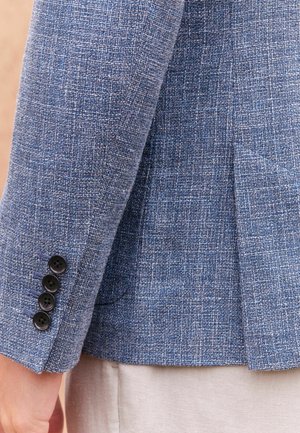 Manica di blazer blu testurizzato con quattro bottoni neri e una spaccatura al polsino, che mette in evidenza il motivo tessuto del tessuto.