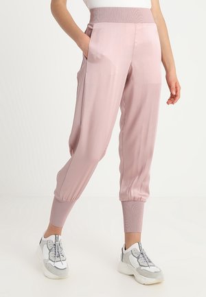 Kobieta nosząca jasnoróżowe, wysokowciskane spodnie zwężane z elastycznymi ściągaczami i kieszeniami, w połączeniu z białymi i szarymi trampkami.