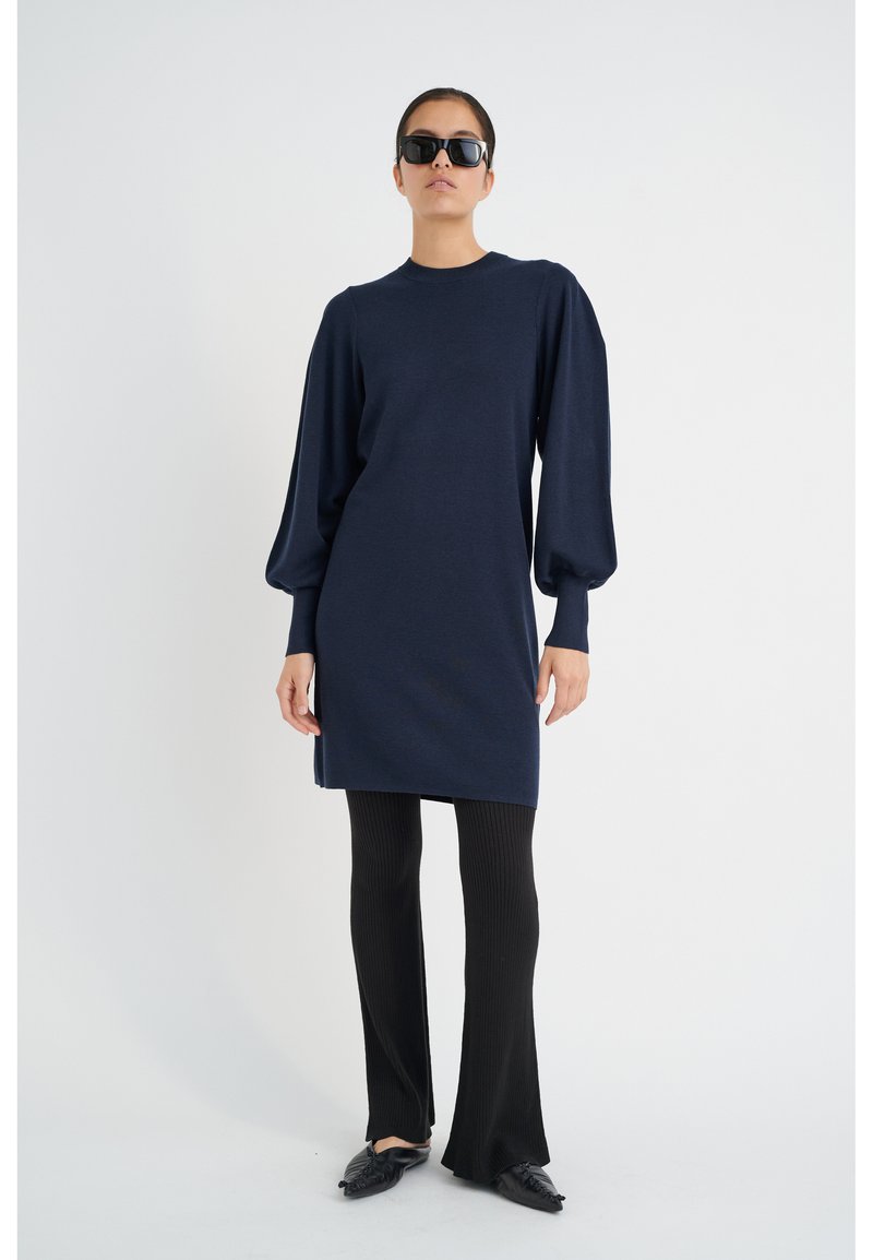Abito tricot blu navy con maniche a sbuffo oversize, scollatura rotonda e taglio dritto, abbinato a pantaloni a zampa neri a coste e scarpe basse.
