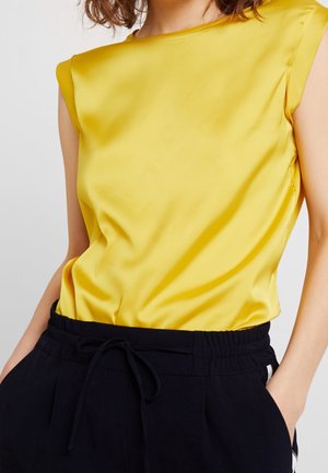 Vrouw draagt een mouwloze gele satijnen top die in zwarte broek met trekkoord is gestopt, met handen in de zakken, zichtbaar van nek tot heupen.