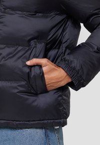 Marine pufferjack met een quiltontwerp, elastische manchetten en een zijzak met een hand erin. Getextureerde, waterbestendige stof.