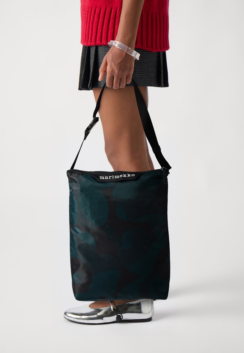 Borsa tote di colore verde scuro con un motivo di foglie, realizzata in tessuto. Presenta manici neri e un'etichetta con il logo nella parte superiore. Tenuta da una persona.