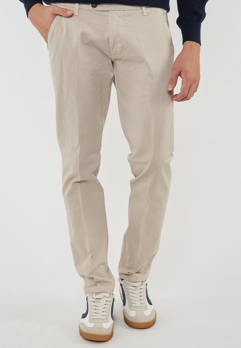 Pantaloni in cotone beige con una vestibilità slim, dotati di zip e tasche. Abbinati a sneaker bianche con dettagli blu navy e suola in gomma.