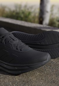 Chaussures de sport noires avec tissu texturé et semelles épaisses en caoutchouc posées sur une surface en béton à l'extérieur, près d'un arrière-plan naturel flou.
