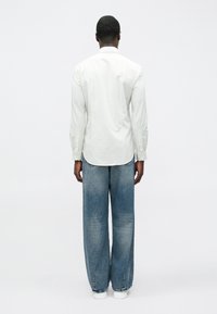 Weißes langärmeliges Hemd mit Streifen und abgerundetem Saum, kombiniert mit hellblauen Jeans mit weitem Bein und weißen Turnschuhen, von hinten gesehen.