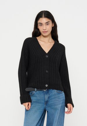 Femme aux longs cheveux foncés portant un pull en maille noir boutonné et un jean bleu sur un fond blanc uni.