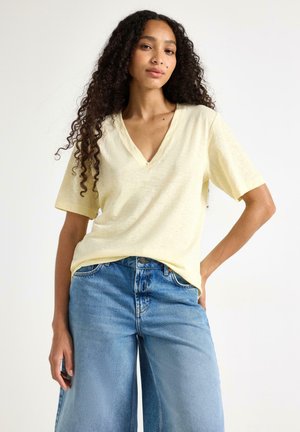 Kvinde med langt krøllet hår iført en lysegul V-hals kortærmet skjorte og højtaljede løse blå jeans, poserer med den ene hånd på hoften.