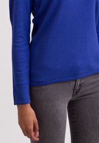 Koninklijk blauwe trui met lange mouwen, gemaakt van zachte gebreide stof, gecombineerd met grijze spijkerbroek. Naaddetails op de manchet en zoom.