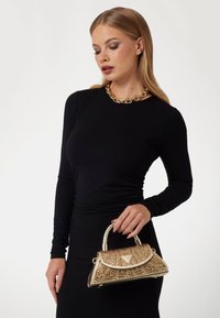 Borsa in oro metallico con intricate perline, manico arrotondato e forma strutturata. Indossata con un vestito nero a maniche lunghe.