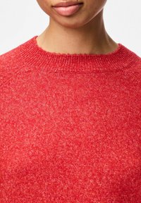 Raudonas megztinis su apvalia iškirpte, tekstūruotas audinys, turintis šiek tiek pūkuotą išvaizdą, ir subtilios spalvų variacijos visame paviršiuje.