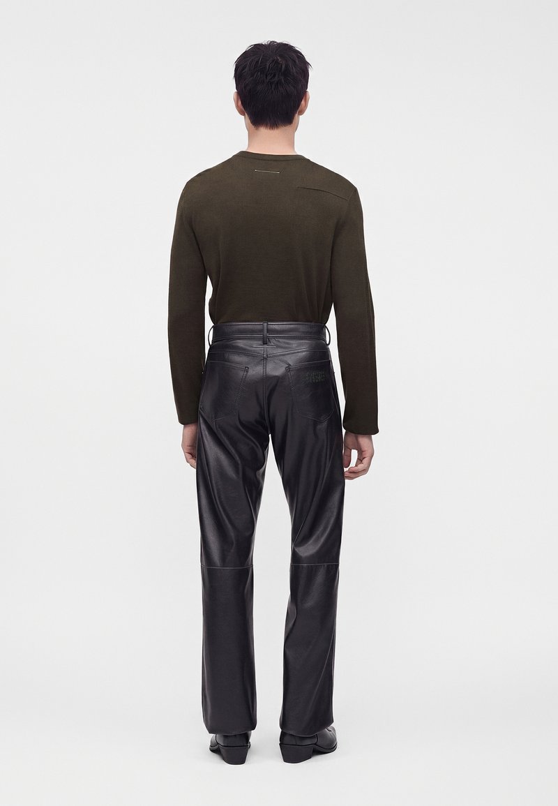 Man wearing an olive long-sleeve shirt with high-waisted black leather pants and black shoes, standing facing away on a plain background.