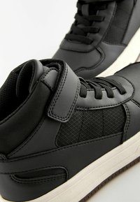 Zwarte hoge sneakers met glad leer en meshpanelen, met platte zwarte veters, een haak-en-lusriem en een witte tussenzool.