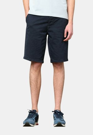 Uomo che indossa pantaloncini blu navy fino al ginocchio, una camicia azzurra e scarpe da ginnastica blu, in piedi con le mani ai lati.