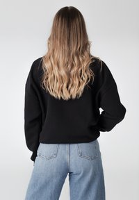 Sweatshirt noir oversize avec épaules tombantes, associé à un jean taille haute bleu clair. Des cheveux ondulés proéminents tombent au-delà des épaules.