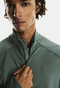 Gaiši zaļa, teksturēta jaka ar rāvējslēdzēju, augstu apkakli un caurumiem īkšķiem; tai ir logo uz krūtīm un rāvējslēdzēja detaļa.