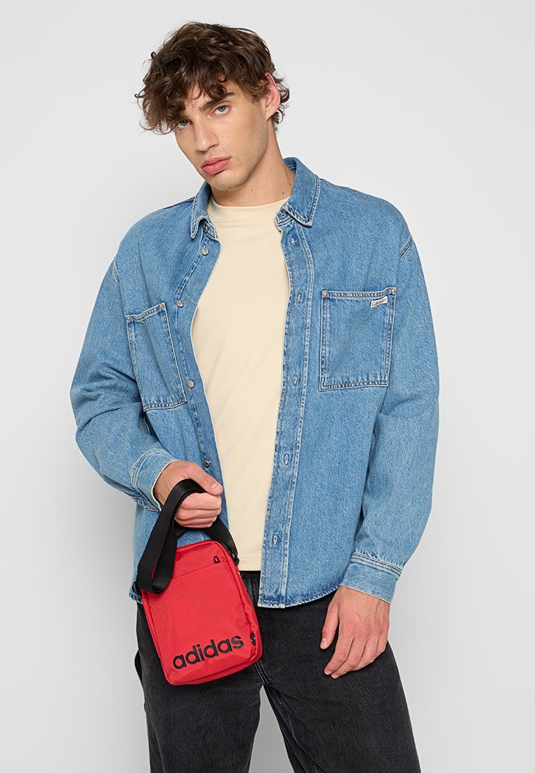 Bolso de hombro rojo de Adidas con logo negro, sostenido por una persona que lleva una camisa de mezclilla azul sobre una camiseta crema y pantalones negros.
