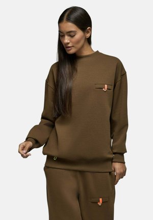 Femme portant un sweat-shirt et un pantalon marron assortis avec de petits onglets orange sur les poches, debout devant un fond uni.