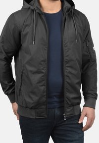 Svart jacka med dragkedja och huva, gjord av mjukt, lätt tyg. Har elastiska muddar och midjeband, framfickor med dragkedja samt detaljering vid sidorna.